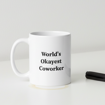 White mug with 'World's Okayest Coworker' text on a white surface with a black pen.